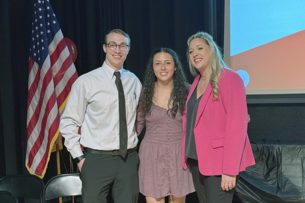 Three people standing together in front of an American flag and a presentation screen at Casper College.
