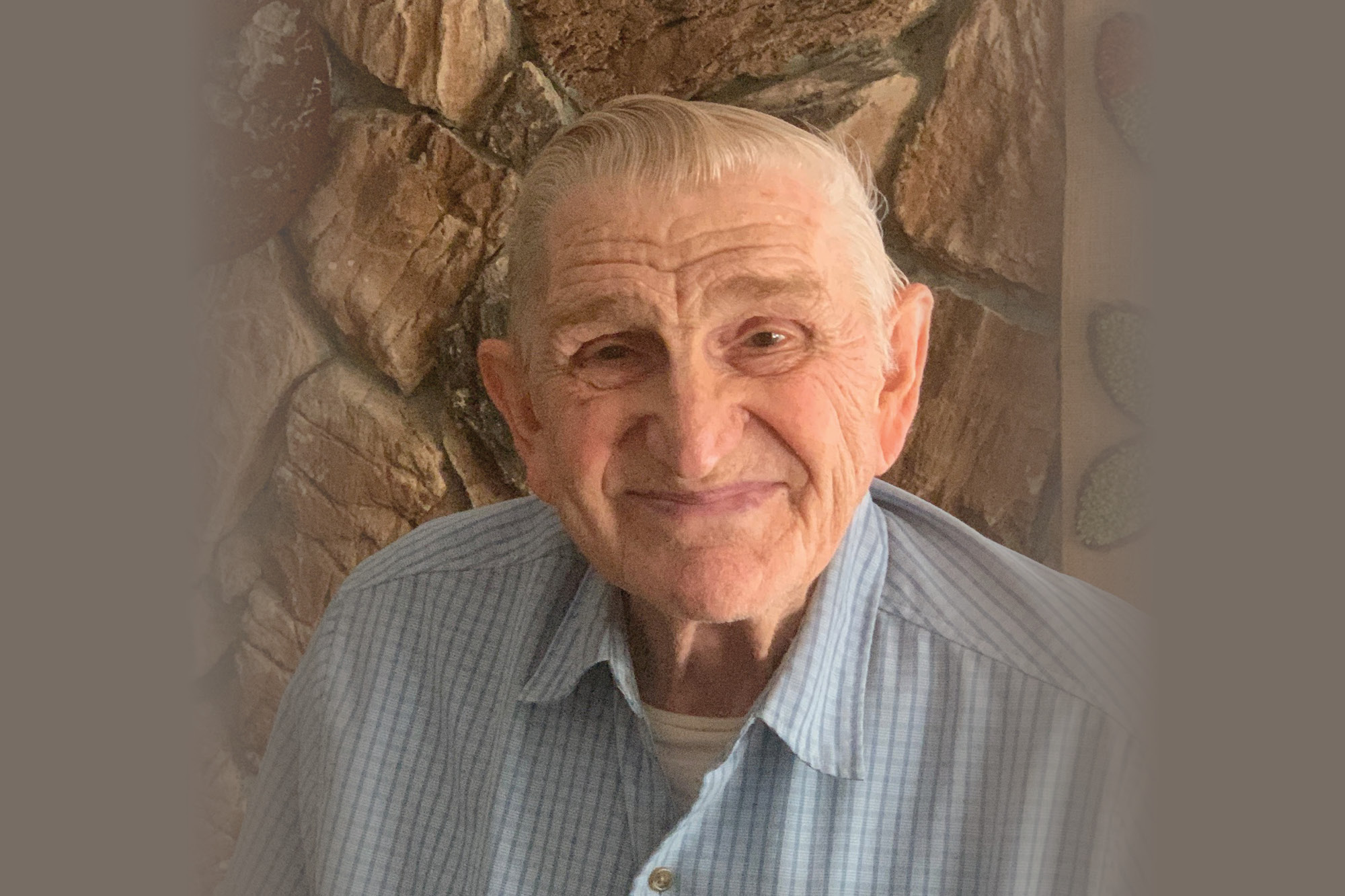 Portrait of Michael Burger, an elderly man with a kind smile wearing a blue striped shirt, standing in front of a stone wall.
