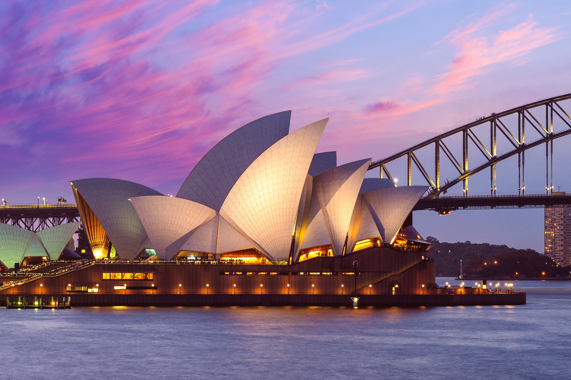 Dusk photograph of the illuminated Sydney Opera House and the Sydney Harbour Bridge, set against a vibrant purple and pink sky.