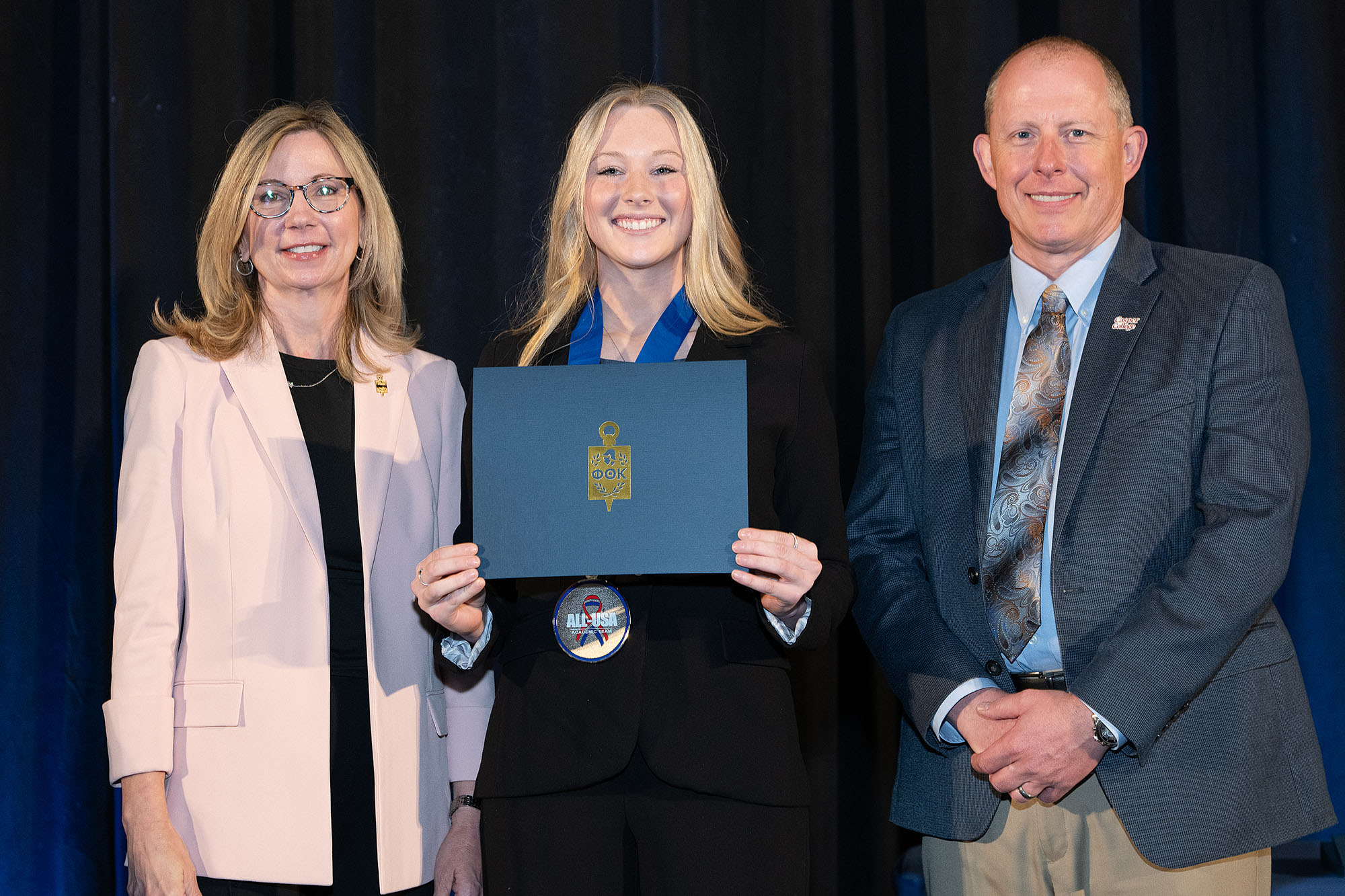A PTK representaive, Ahnya Ivie-Moody, and Dr. Brandon Kosine holding a Phi Theta Kappa certificate and wearing an All-USA Academic Team medal.