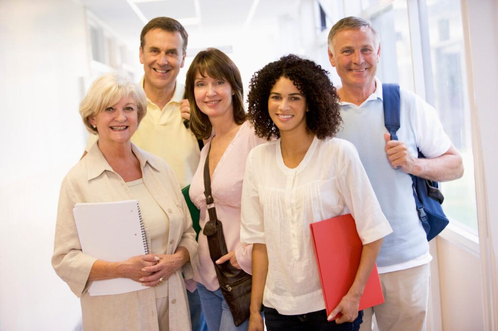 Five smiling adult students standing in a brightly lit hallway holding notebooks and folders for community education classes